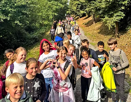 Schüler:innen wandern durch einen bewaldeten Weg und winken in die Kamera