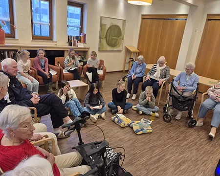Kinder sitzen auf dem Boden während ältere Menschen im Kreis sitzen. Ein Mädchen liest etwas vor